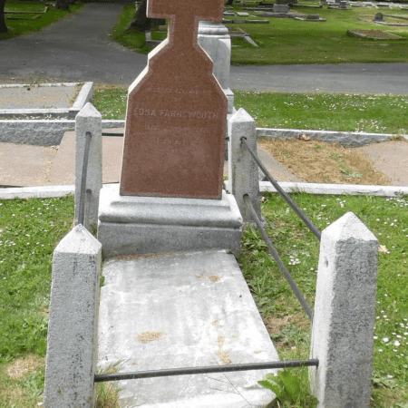 A tilted gravestone with a cross stands behind a sunken and shifted stone slab, bordered by four short stone posts connected by metal rods, in a grassy cemetery with other headstones and trees in the background.