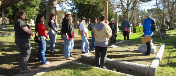Group of university students and a professor participating in an archaeology class tour at Ross Bay Cemetery, discussing graves and monuments in a sunny outdoor setting.